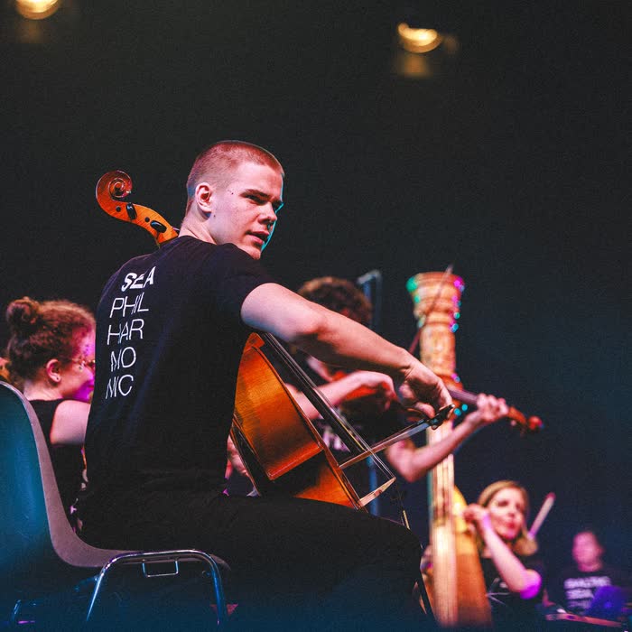 Kaur Nellis playing cello in a Baltic Sea Philharmonic shirt, looking back over his shoulder mid-performance.