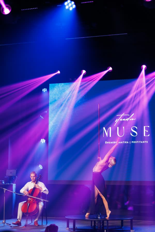 Kaur Nellis playing cello at the left of a dark stage while aerialist Lisann Lumet performs on a vertical pole at the right, under magenta and blue beams.