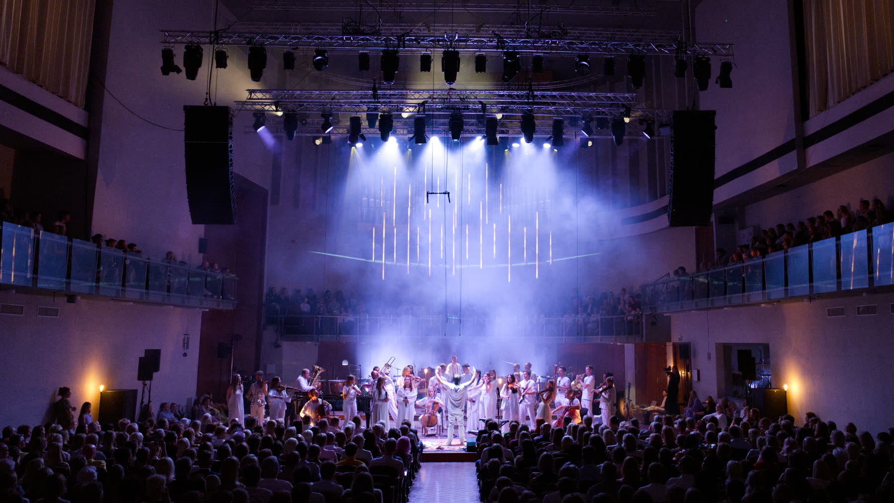A packed concert hall, the ensemble in white on a hazy stage lit by vertical light tubes.